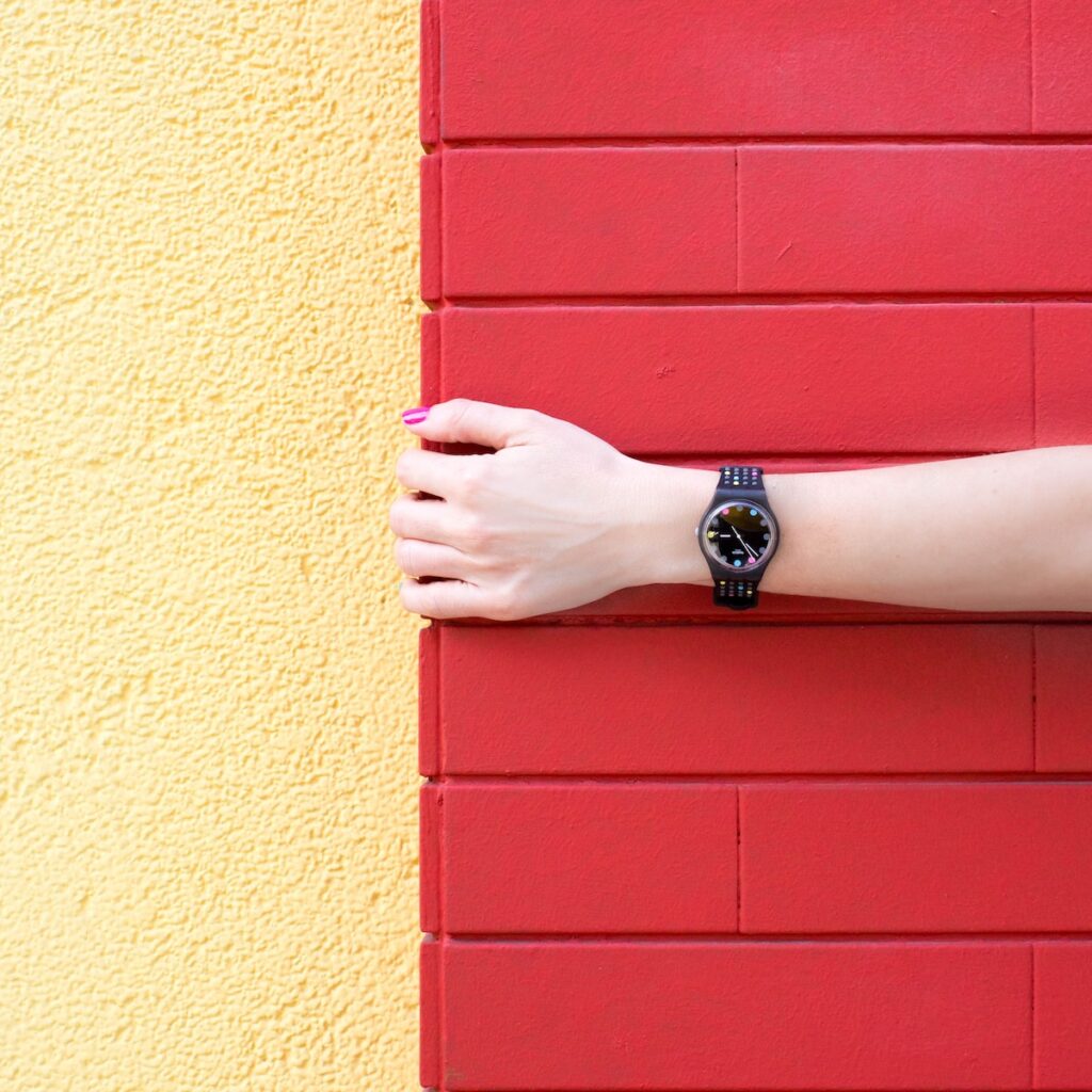 Swatch watch on wrist with a lot of color in background person wearing black analog watch holding red brick wall